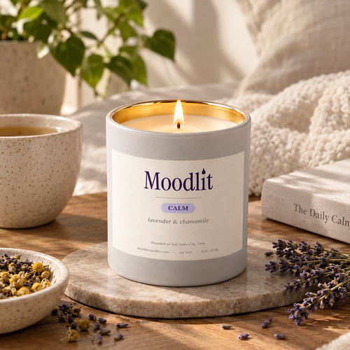 A lit Moodlit candle in a light gray jar with a gold inner rim sits on a wooden surface in a cozy styled setting with a mug, books, dried flowers, and soft fabric in the background.