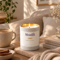 A lit Moodlit Cozy candle in a white glass jar with a gold rim sits on a wooden table in a cozy living room setting, surrounded by a ceramic mug, cinnamon sticks, books, and a knitted blanket.