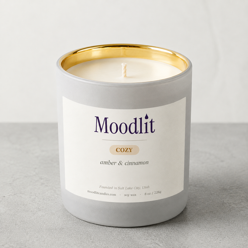A Moodlit Cozy soy wax candle in a matte gray jar with a gold interior rim, cream-colored wax, and a white label reading 'Moodlit COZY amber & cinnamon' on a light gray surface.