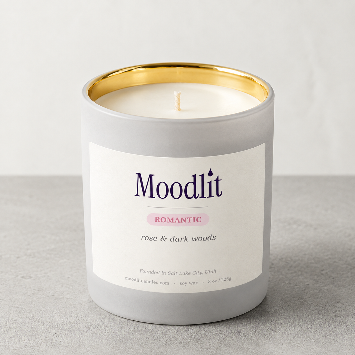 A Moodlit 'Romantic' soy wax candle in a matte gray jar with a gold inner rim and a white label, set on a gray surface against a light gray background.